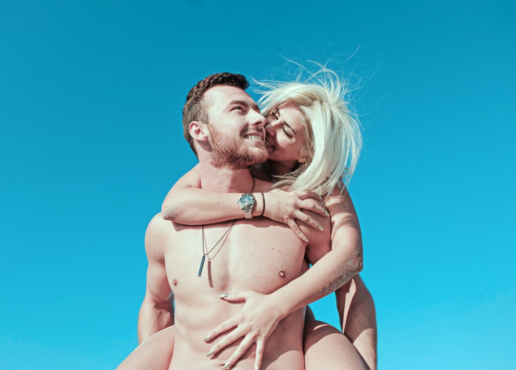 A woman riding on her boyfriend's back at the beach kissing his cheek