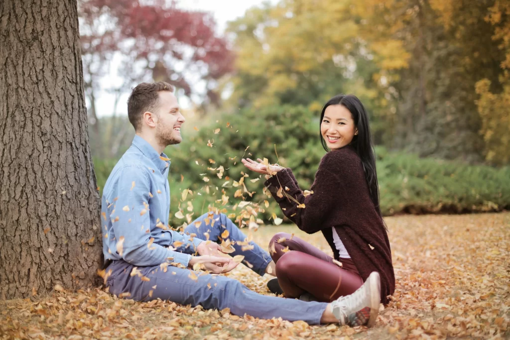 Man and asian woman in a park tossing leaves while sitting beside a tree